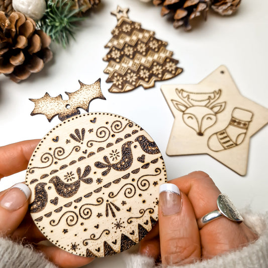 A hand holds a round pyrographed wooden Christmas ornament with intricate holiday patterns, with other pyrography wooden ornaments and pinecones in the background.
