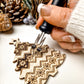 A close-up of a hand using a black-handled pyrography tool to create geometric and floral patterns on a wooden Christmas tree ornament, with pinecones in the background.