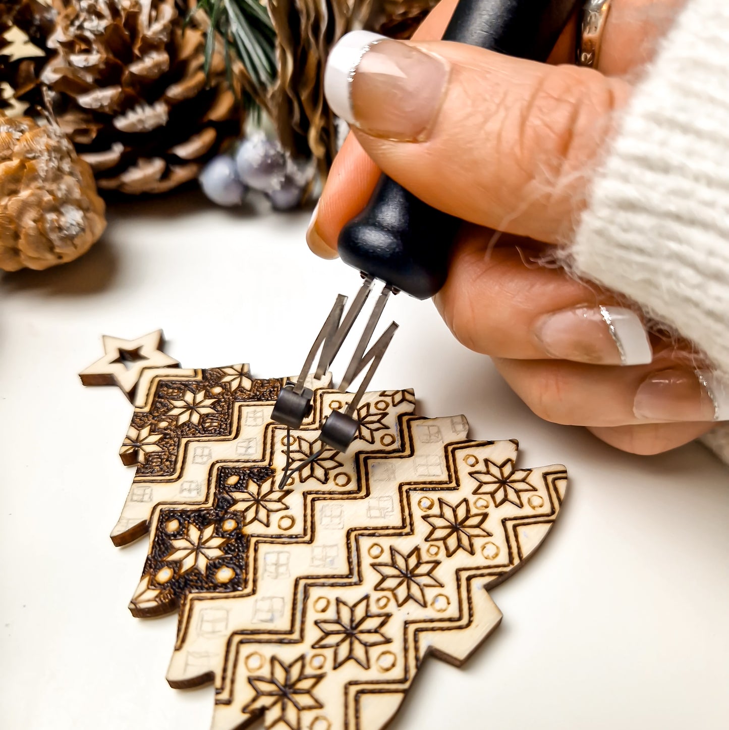 A close-up of a hand using a black-handled pyrography tool to create geometric and floral patterns on a wooden Christmas tree ornament, with pinecones in the background.