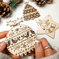 A hand holds a round pyrographed wooden Christmas ornament with intricate holiday patterns, with other pyrography wooden ornaments and pinecones in the background.