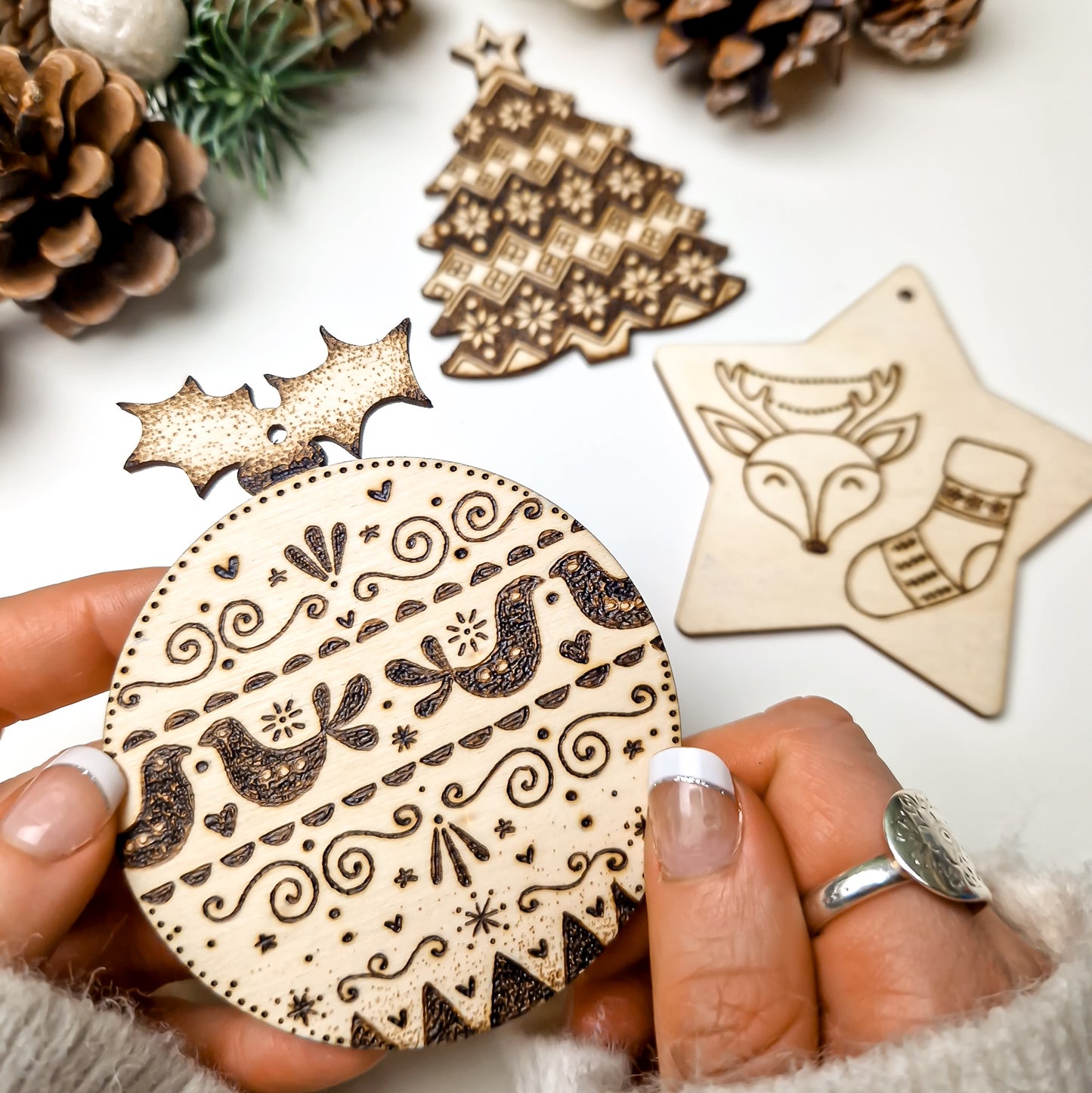 A hand holds a round pyrographed wooden Christmas ornament with intricate holiday patterns, with other pyrography wooden ornaments and pinecones in the background.