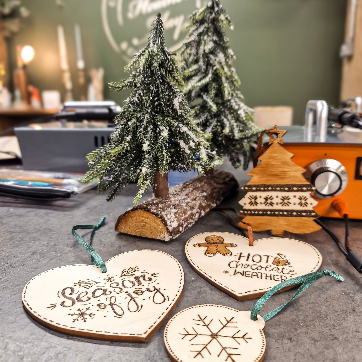 Three pyrography wooden Christmas ornaments shaped like hearts, a gingerbread man, and a snowflake, displayed on a tabletop with miniature Christmas trees and a rustic log in the background.