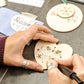A close-up of hands using a Peter Child pyrography tool to burn designs onto a light beige wooden ornament. Other wooden ornaments with burned text and snowflakes are visible on a dark gray tabletop.