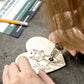 A close-up of a person using a wood burning tool to burn a fox design onto a heart-shaped wooden decoration, with hands and hair visible against a dark textured tabletop.