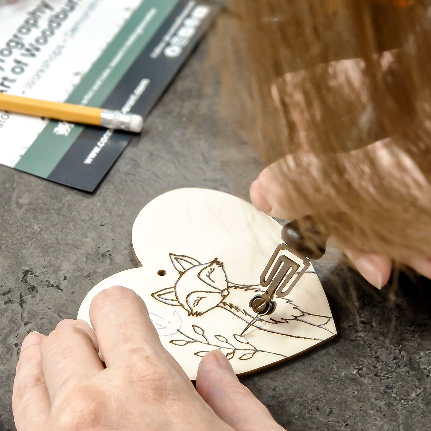 A close-up of a person using a wood burning tool to burn a fox design onto a heart-shaped wooden decoration, with hands and hair visible against a dark textured tabletop.