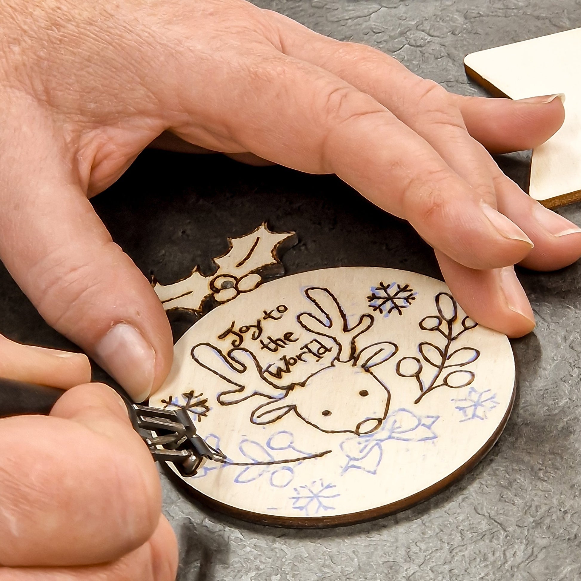 Close-up of a person's hands using a pyrography tool to create a personalised wooden ornament with a reindeer design and the text 'Joy to the world'